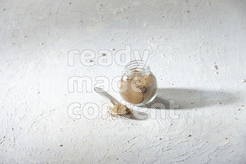 A glass spice jar and metal spoon full of cumin powder on textured white flooring