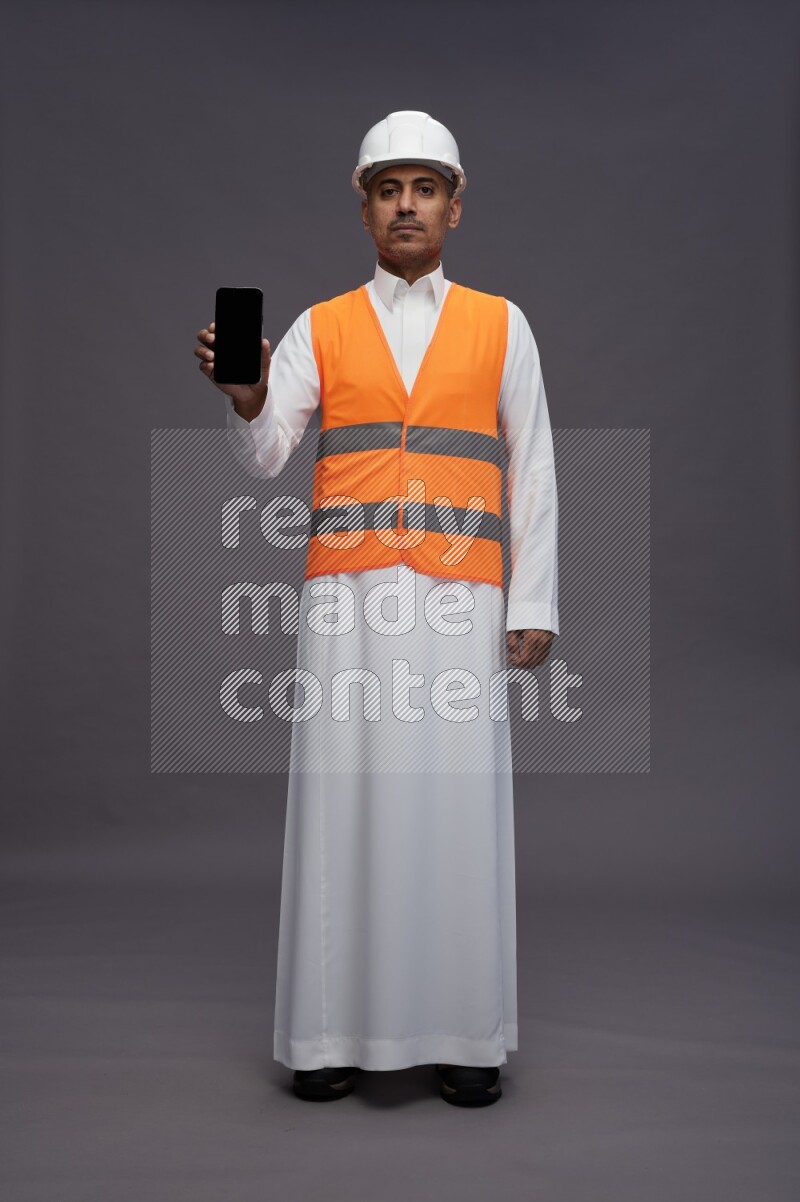 Saudi man wearing thob with engineer vest standing showing phone to camera on gray background