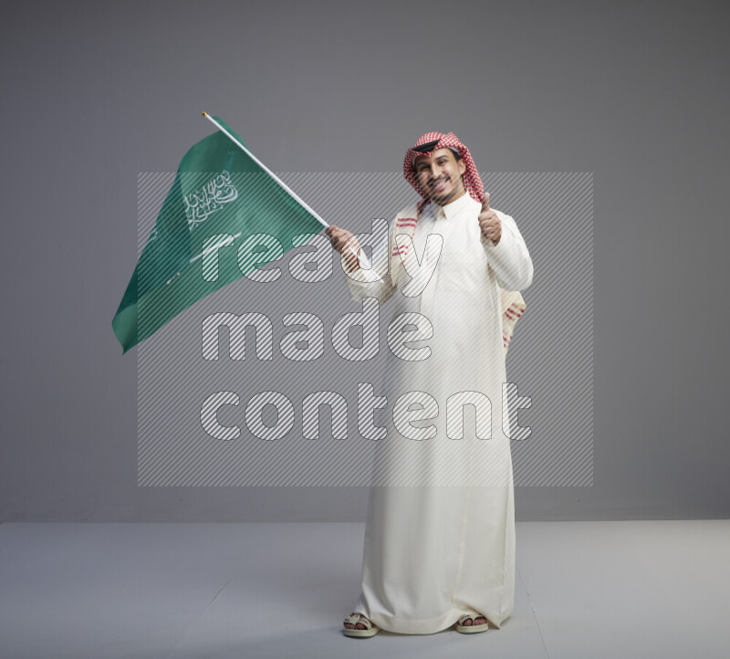 A Saudi man standing wearing thob and red shomag raising big Saudi flag on gray background