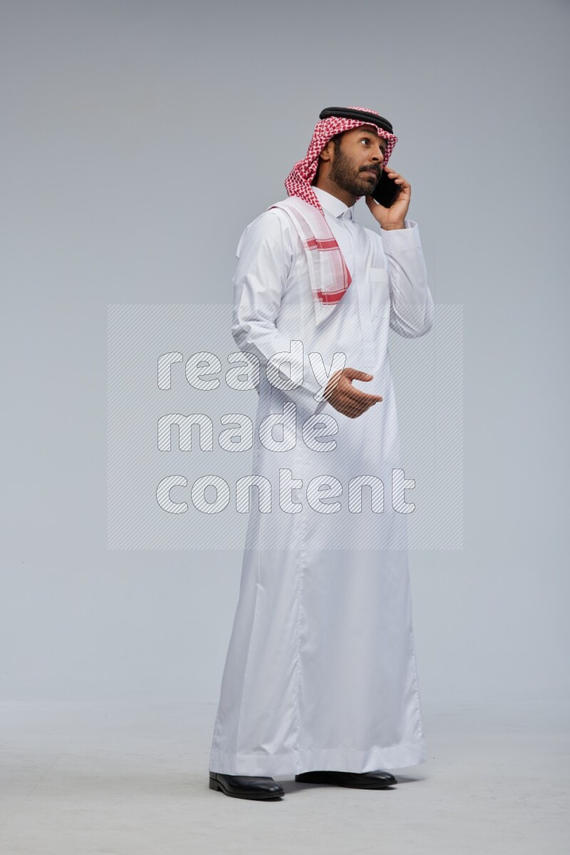 Saudi man Wearing Thob and shomag standing talking on phone on Gray background