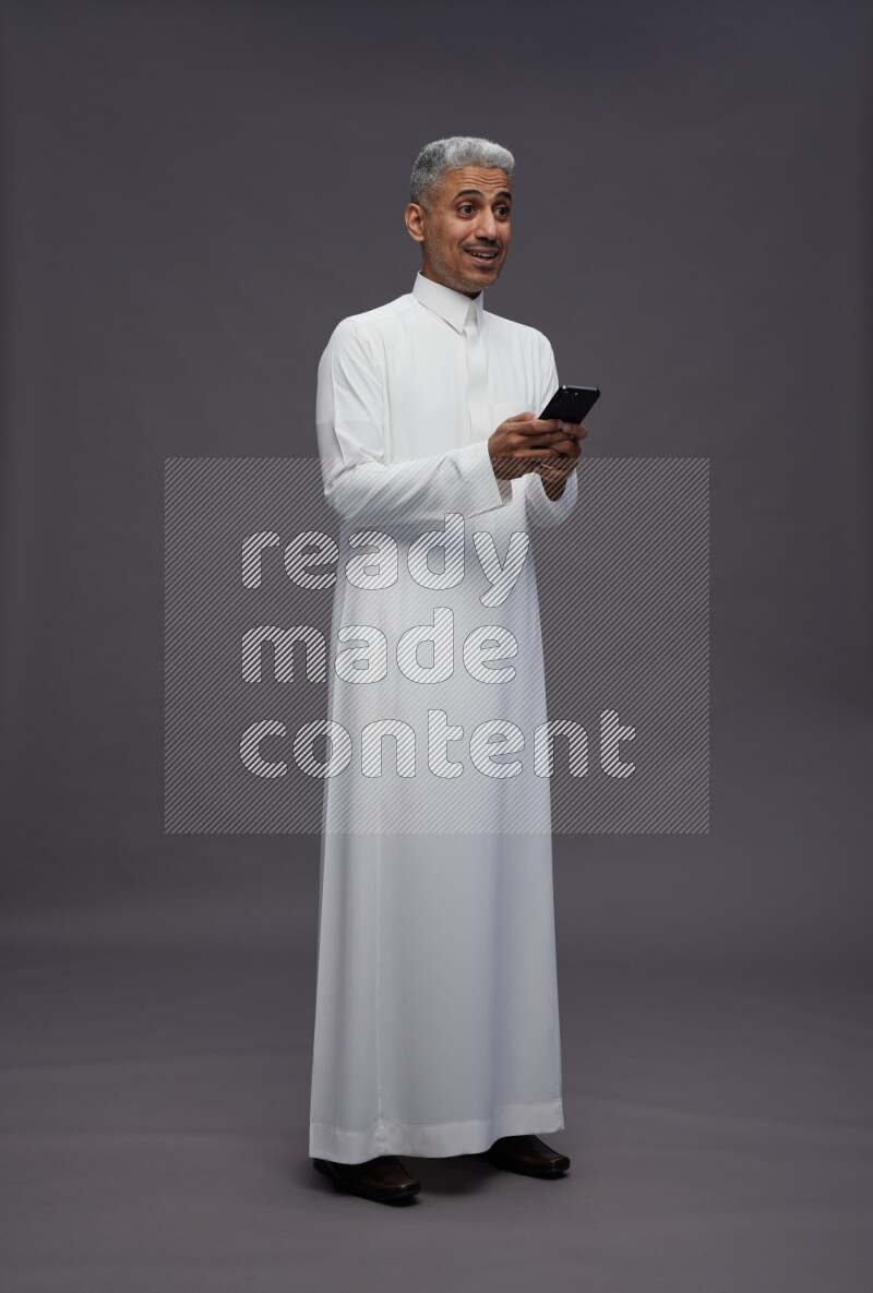 Saudi man wearing thob standing texting on phone on gray background