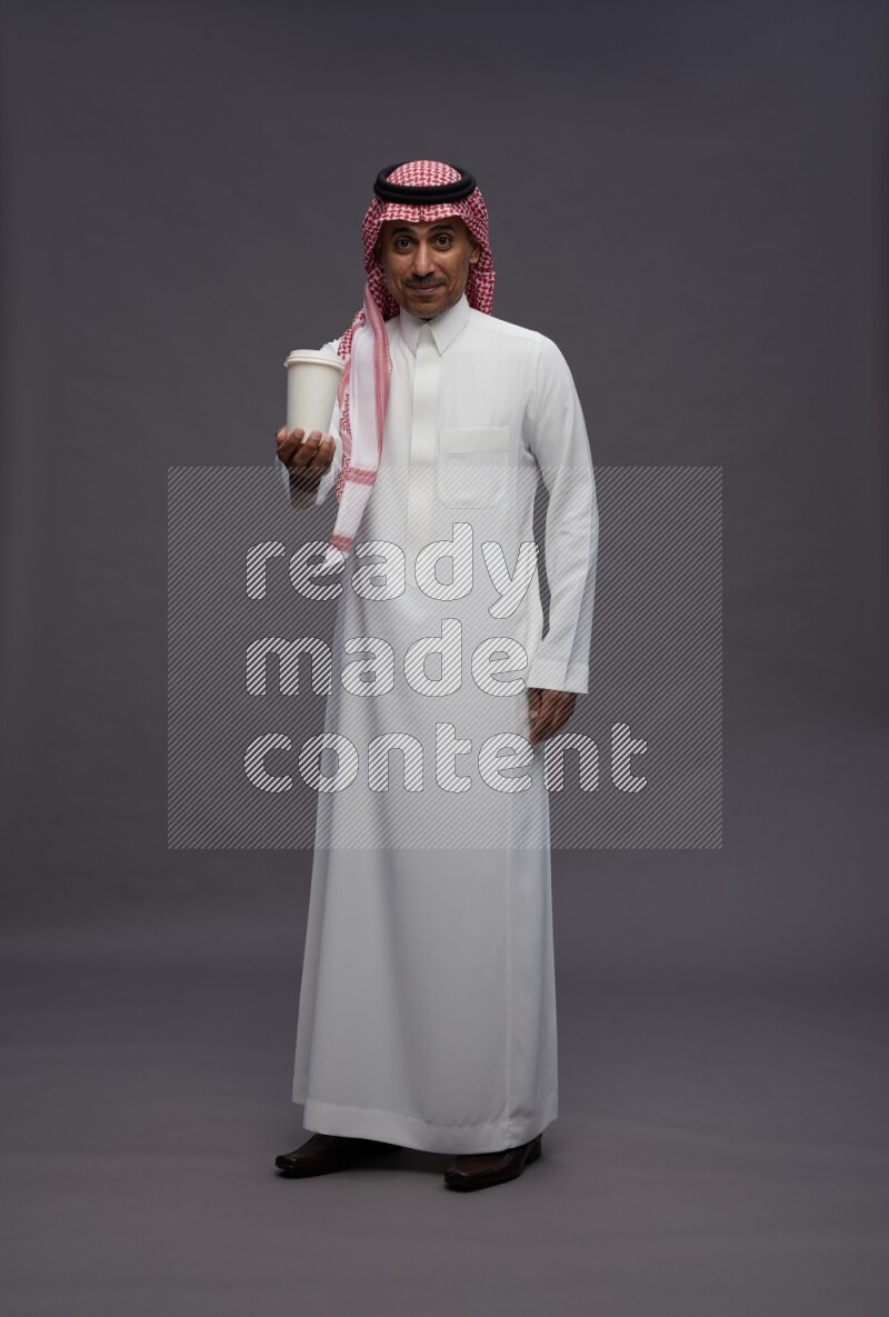 Saudi man wearing thob and shomag standing holding paper cup on gray background