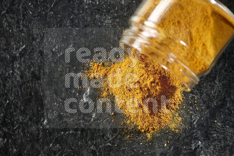 A flipped glass jar full of turmeric powder and powder spilled out of it on a textured black flooring