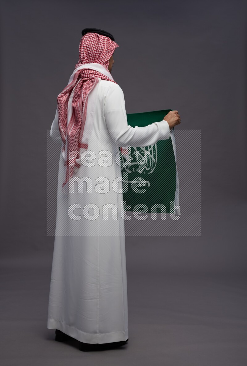 Saudi man wearing thob and shomag standing holding Saudi flag on gray background