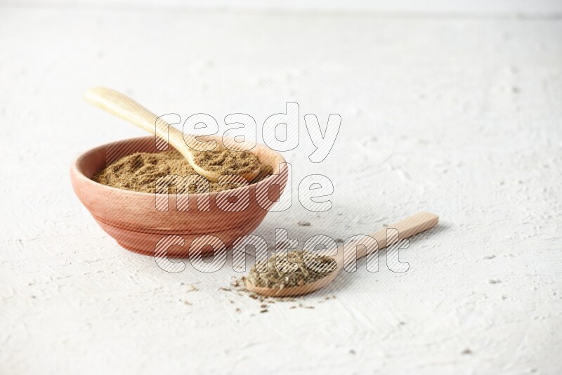 A wooden bowl and 2 wooden spoons full of cumin powder and cumin seeds on textured white flooring