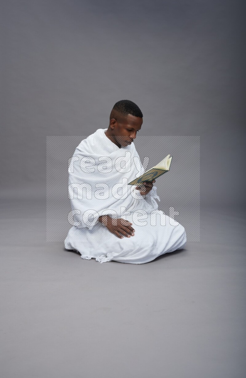 A man wearing Ehram sitting on floor reading quran on gray background