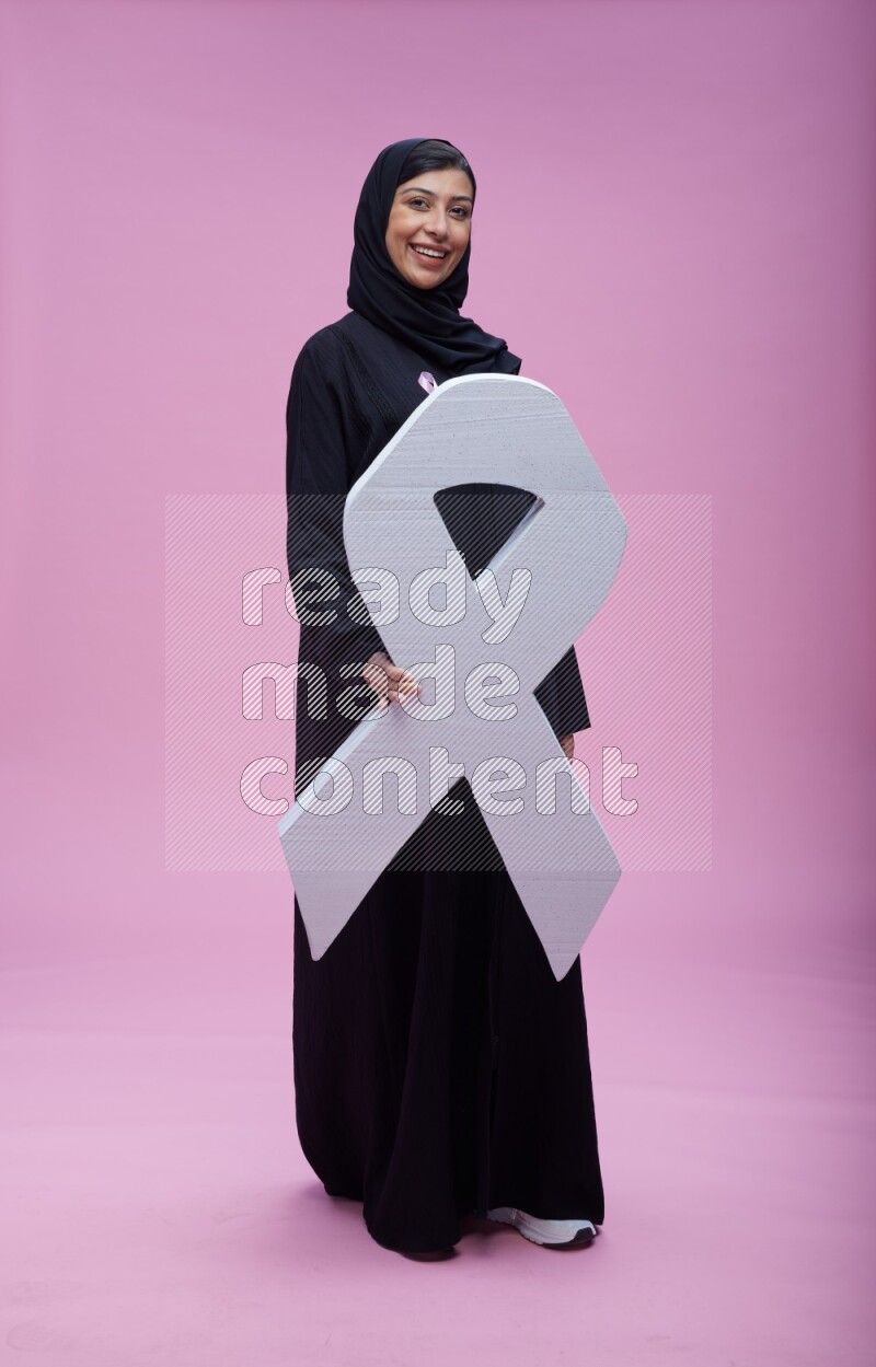 Saudi woman wearing pink ribbon on abaya standing holding awareness ribbon on plain pink background