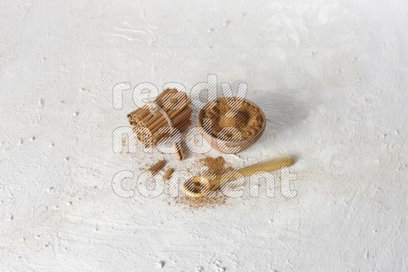 Cinnamon sticks stacked and bounded beside a wooden bowl full of cinnamon powder and a wooden spoon full of powder on white background