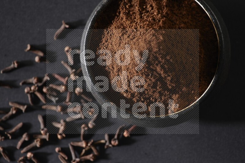 A black pottery bowl full of cloves powder and cloves whole grains on a black flooring