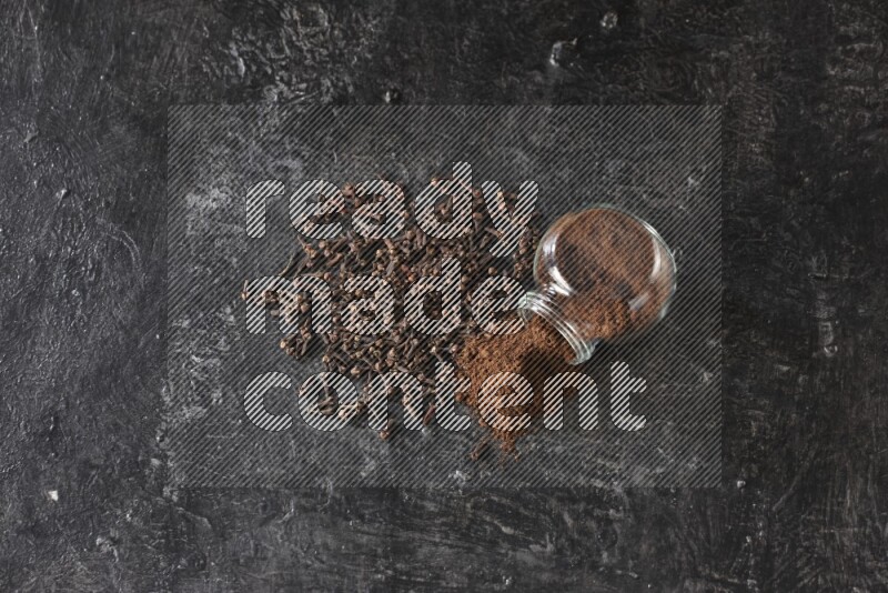 A flipped glass spice jar full of cloves powder and powder came out of it with cloves spread on textured black flooring