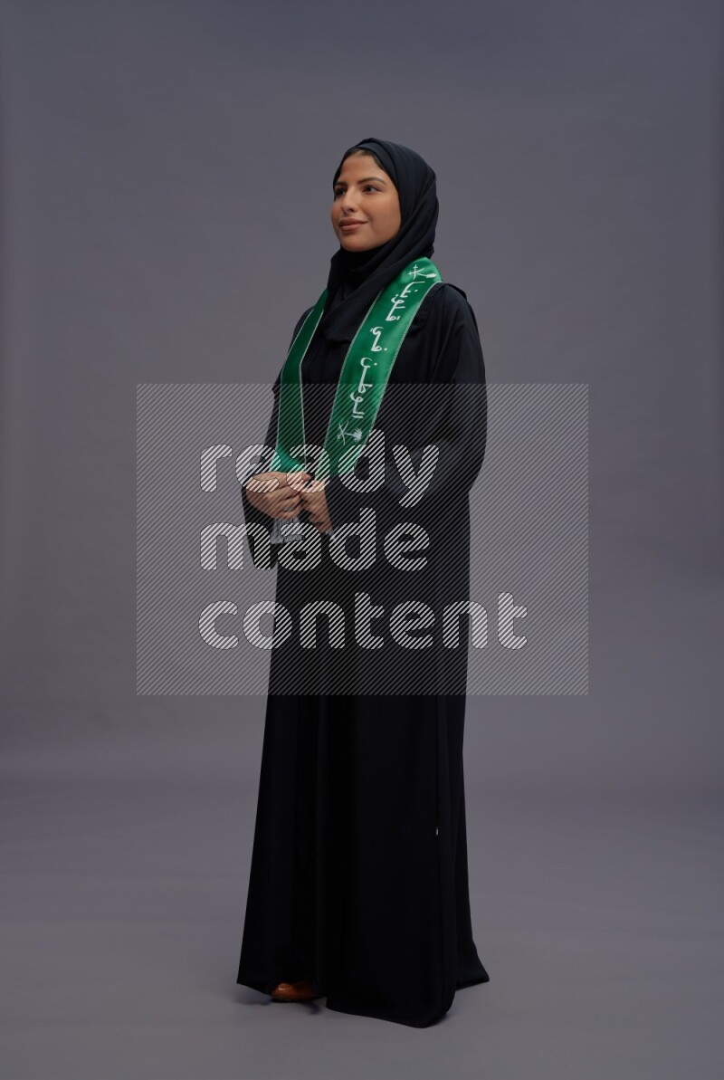 Saudi woman wearing Abaya standing holding Saudi flag on gray background
