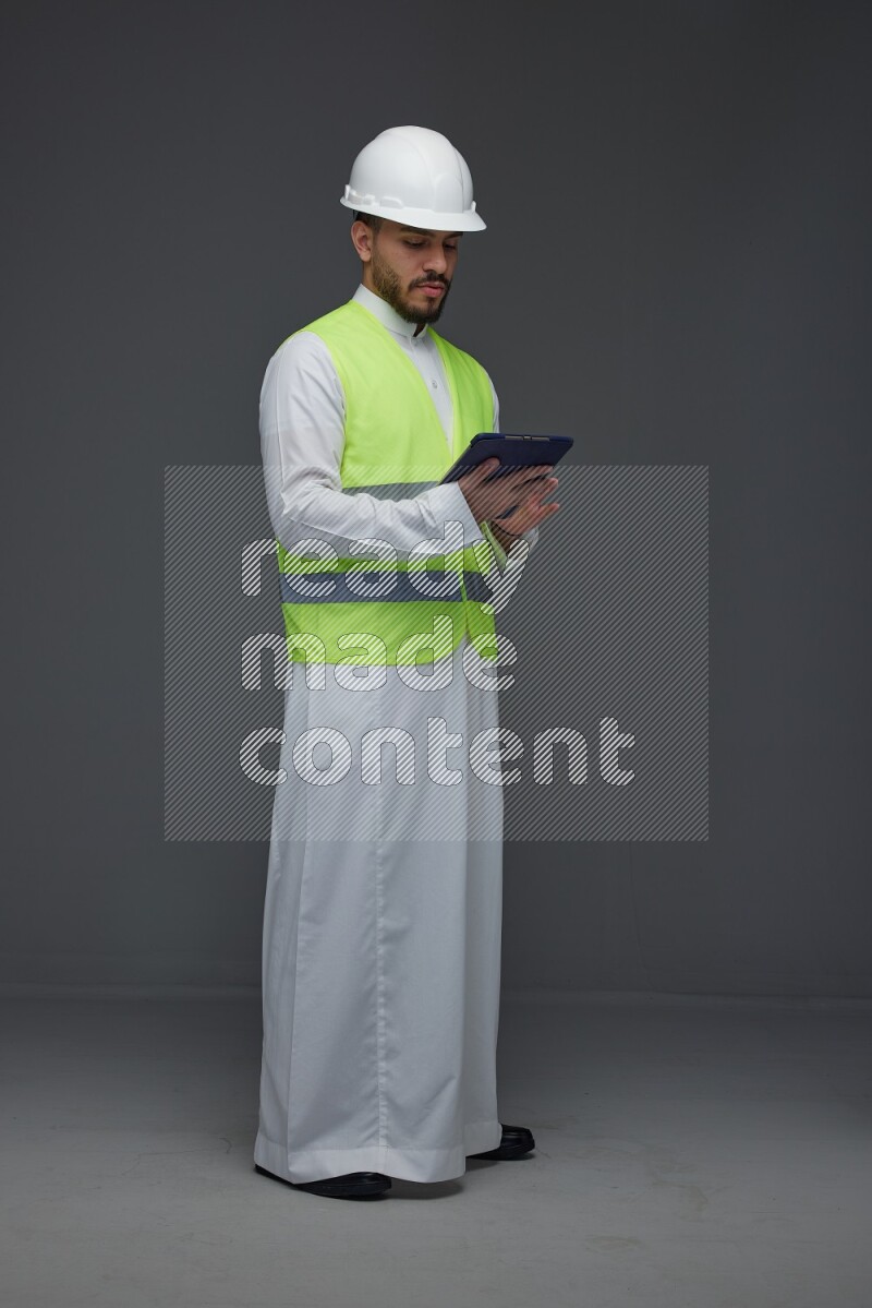 A Saudi man wearing Thobe with a yellow safety vest and white helmet standing and using his tablet different angles eye level on a gray background