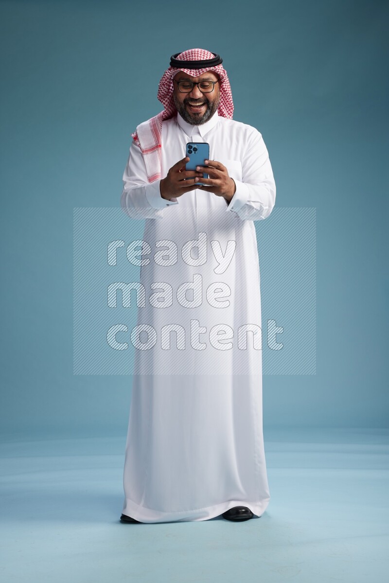 Saudi Man with shimag Standing texting on phone on blue background