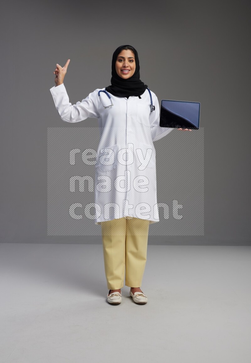 Saudi woman wearing lab coat with stethoscope standing showing tablet to camera with sign in the back on Gray background