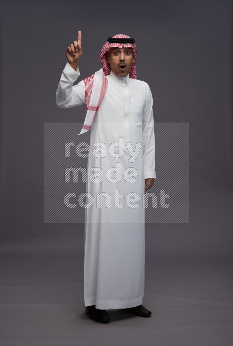 Saudi man wearing thob and shomag standing interacting with the camera on gray background