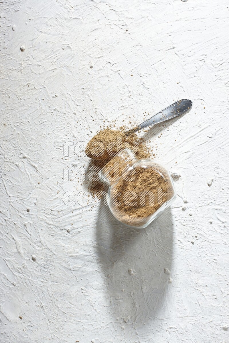 A flipped glass spice jar and a metal spoon full of cumin powder and powder spilled out on textured white flooring