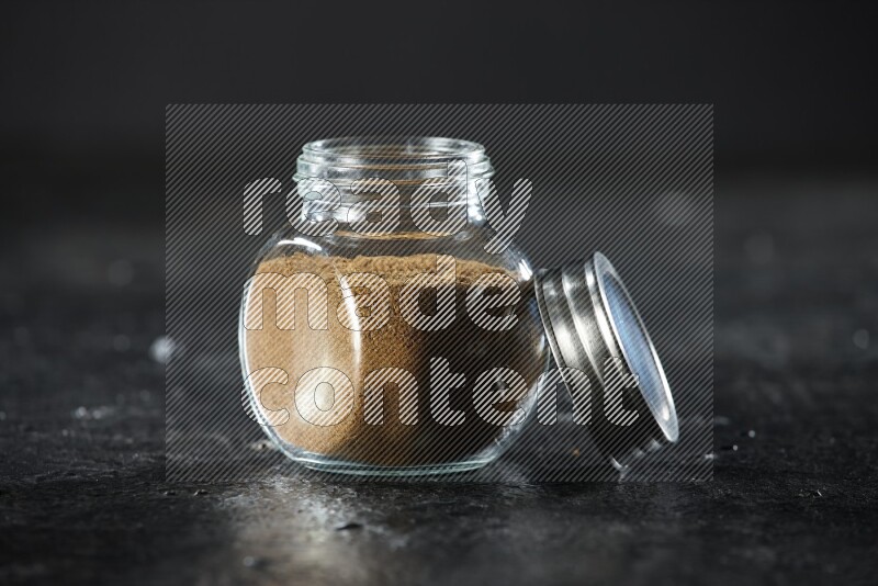 A glass spice jar full of cumin powder on a textured black flooring