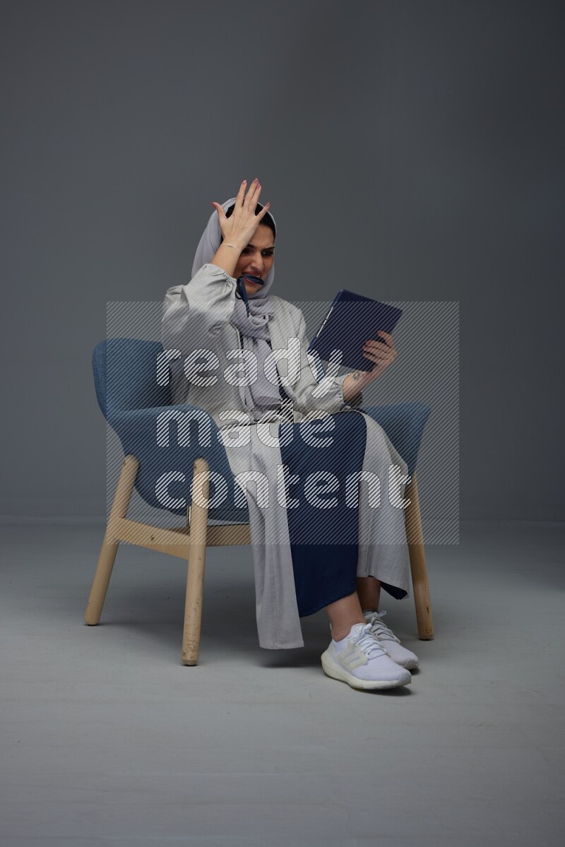 A Saudi female wearing a light gray Abaya and head scarf sitting on a dark grey chair while holding her iPad on a grey background