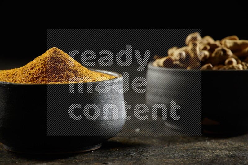 2 pottery black bowls, one full of turmeric powder and the other full of dried turmeric whole fingers on a textured black flooring