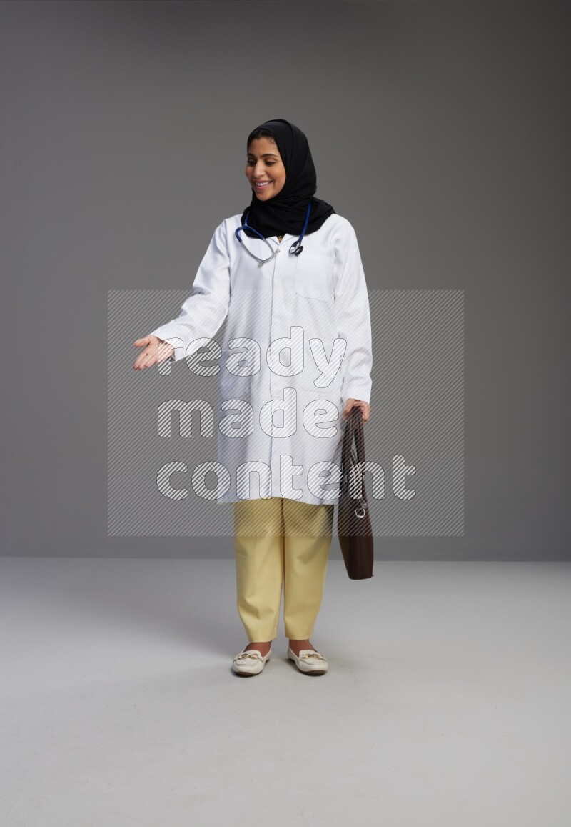 Saudi woman wearing lab coat with stethoscope standing holding bag on Gray background