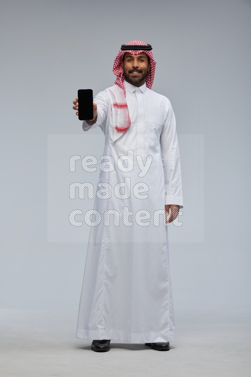 Saudi man Wearing Thob and shomag standing showing phone to camera on Gray background