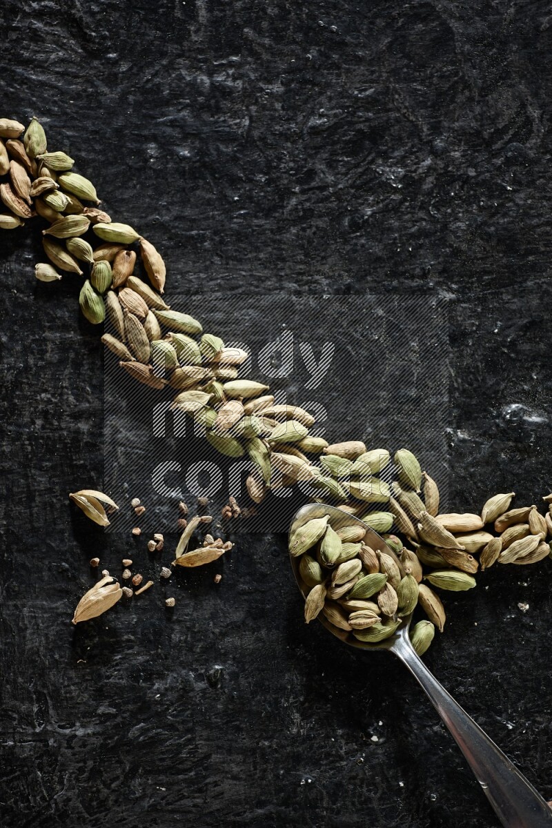 A Metal spoon full of cardamom seeds and some seeds beside it on a textured black flooring