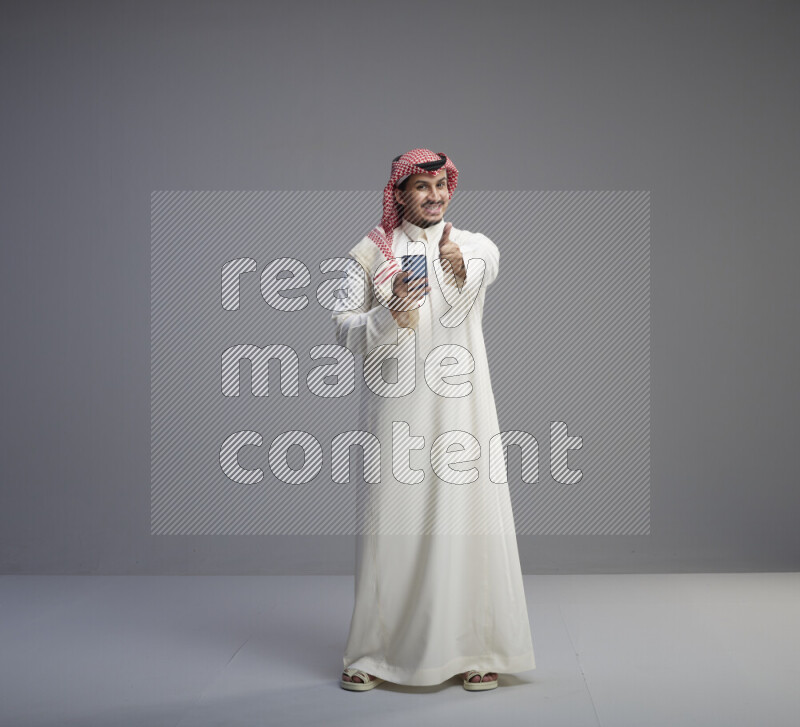 A  Saudi man standing wearing thob and red shomag texting on phone on gray background