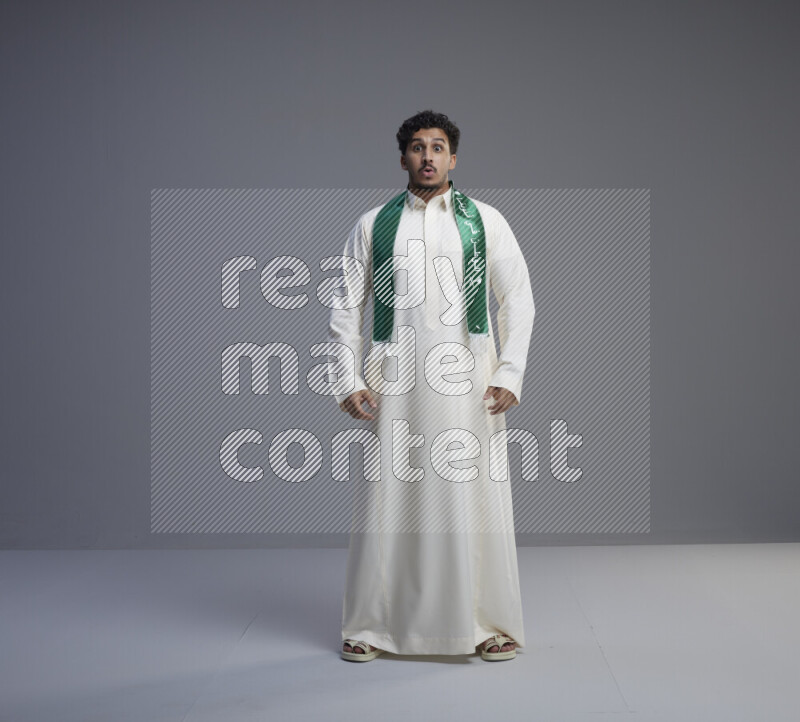 A Saudi man standing wearing thob and Saudi flag scarf on gray background