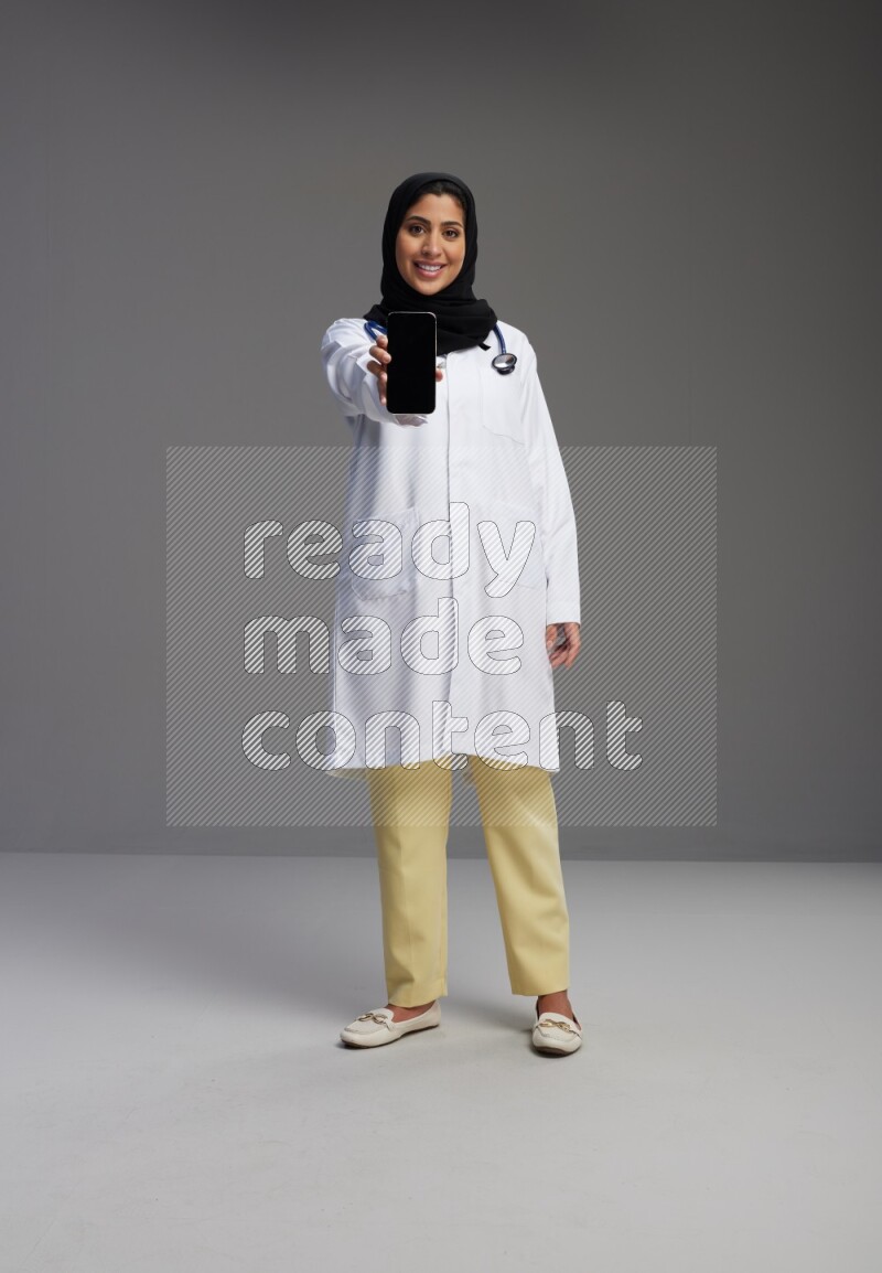 Saudi woman wearing lab coat with stethoscope standing showing phone to camera with sign in the back on Gray background