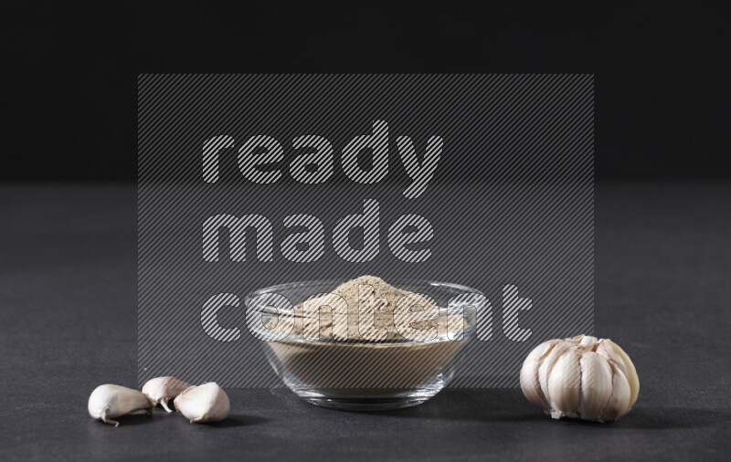 A glass bowl full of garlic powder with garlic bulb and some cloves beside it on a black flooring