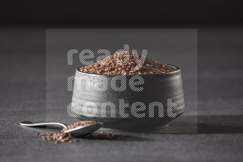 وعاء فخاري أسود ممتلئ بحبوب بذر الكتان مع ملعقة معدنية ممتلئة على أرضية سوداء