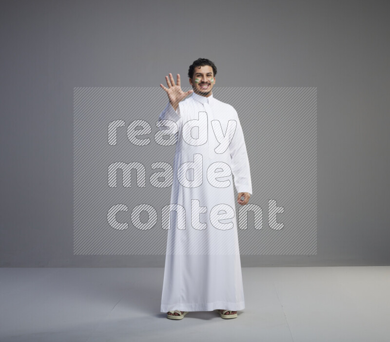 A Saudi man standing wearing thob with face painting interacting with the camera on gray background