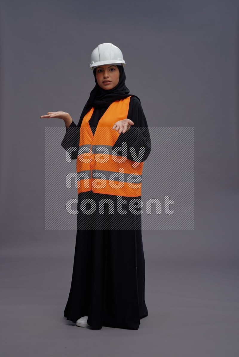 Saudi woman wearing Abaya with engineer vest standing interacting with the camera on gray background