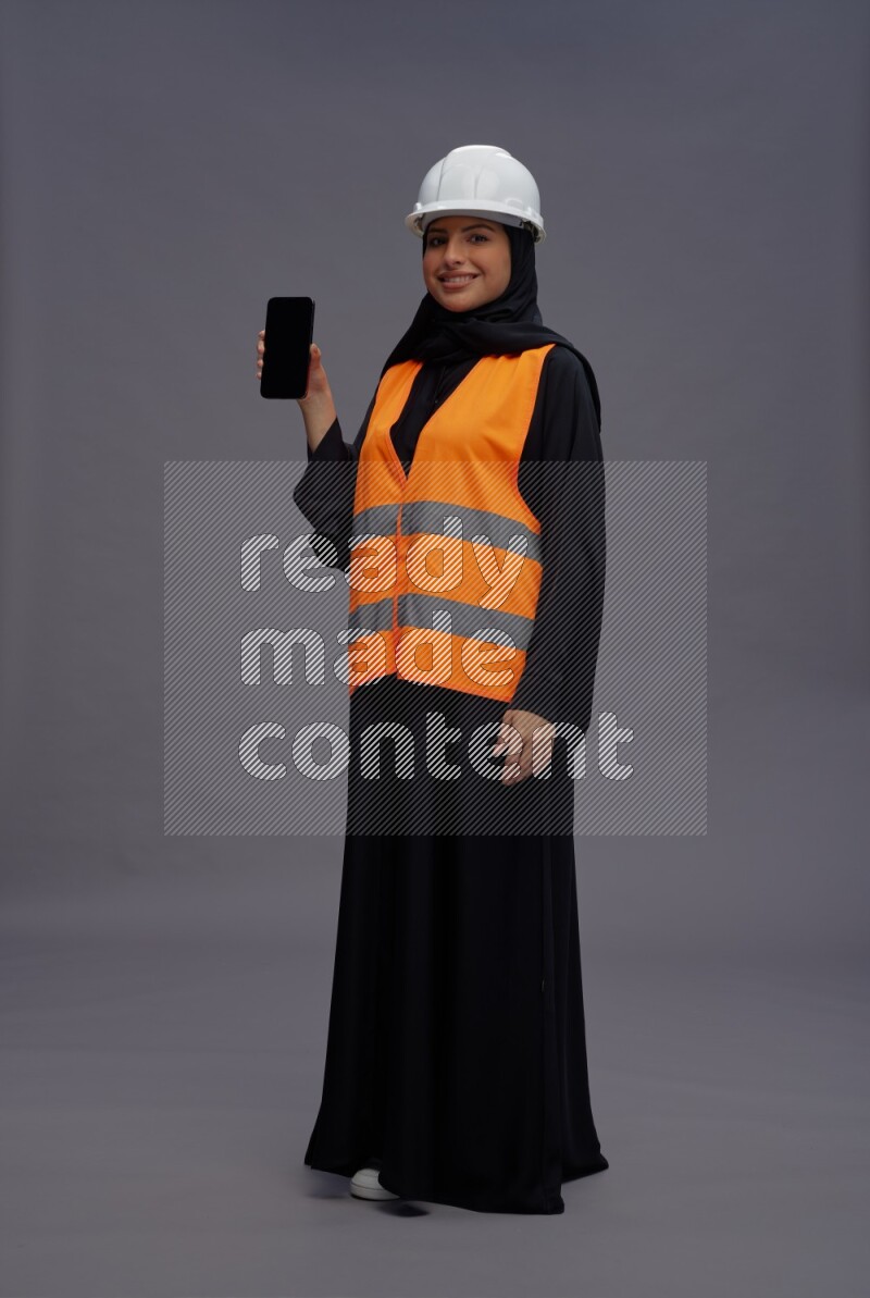 Saudi woman wearing Abaya with engineer vest standing showing phone to camera on gray background