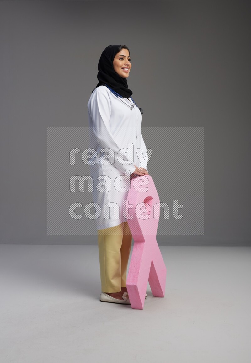 Saudi woman wearing lab coat with stethoscope standing holding awareness ribbon symbol standing on Gray background