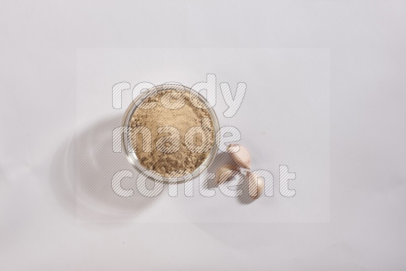 A glass bowl full of garlic powder with garlic bulb and some cloves beside it on a white flooring