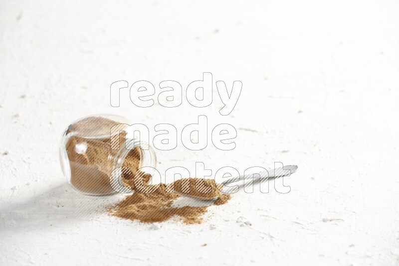 Flipped herbs glass jar full of cinnamon powder with a metal spoon full of powder on a textured white background