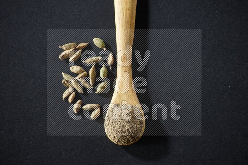 A wooden spoon full of cardamom powder and cardamom seeds beside it on black flooring