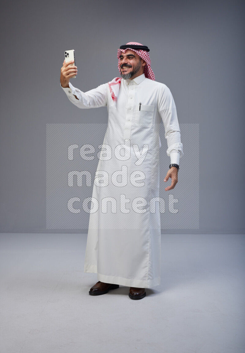 Saudi man Wearing Thob and red Shomag standing taking selfie on Gray background
