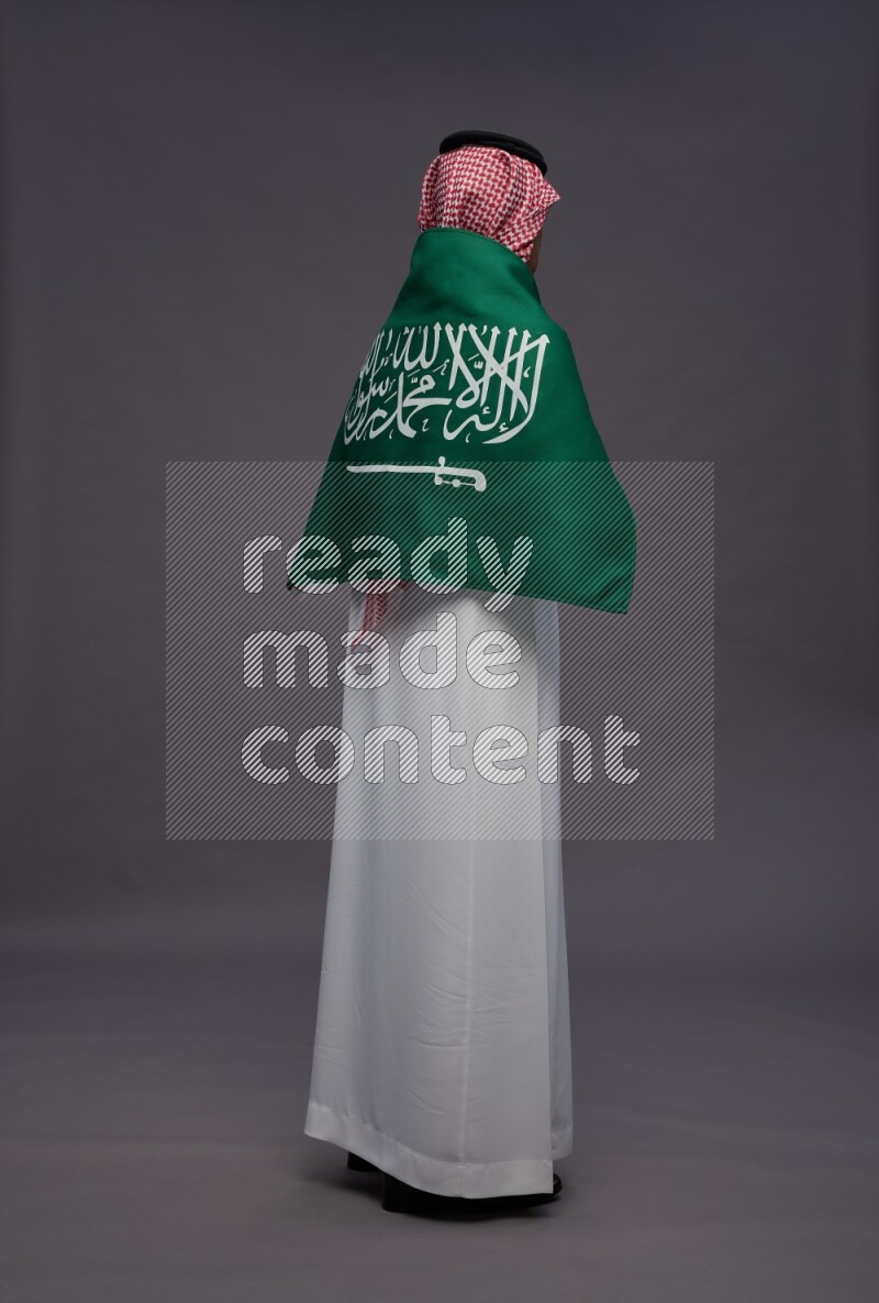 Saudi man wearing thob and shomag standing holding Saudi flag on gray background