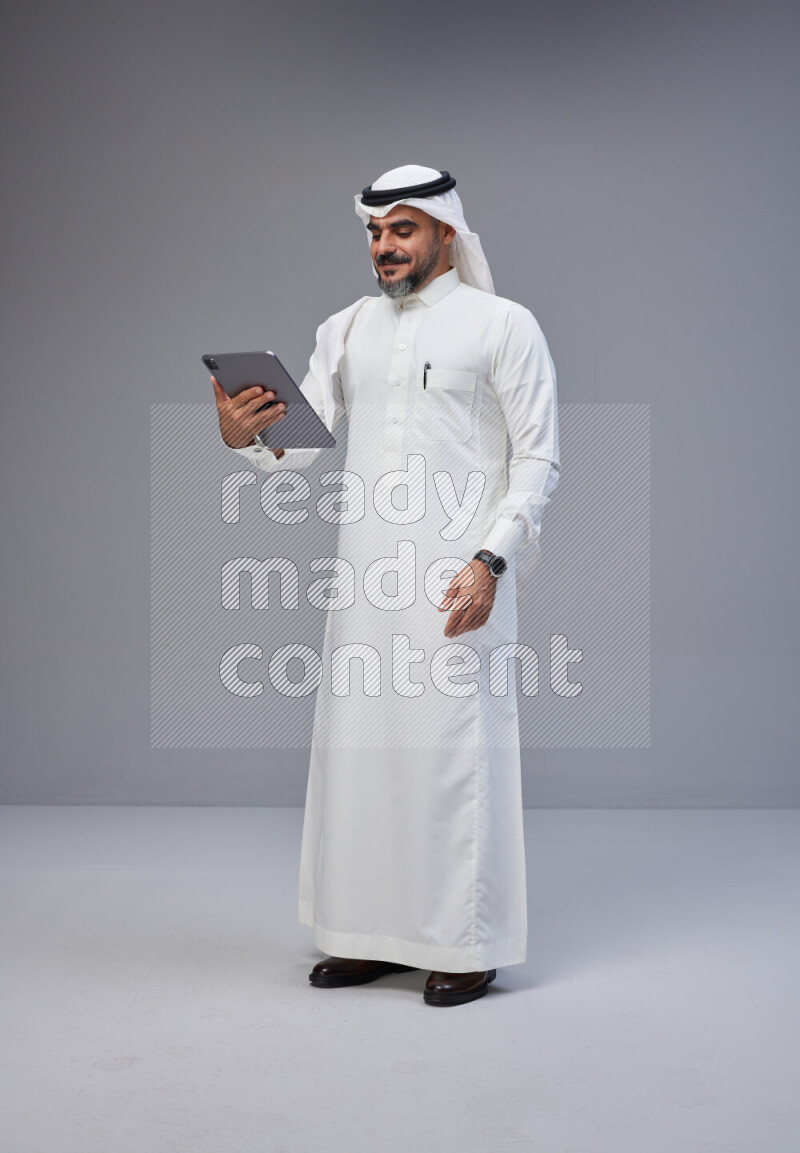 Saudi man Wearing Thob and white Shomag standing working on tablet on Gray background