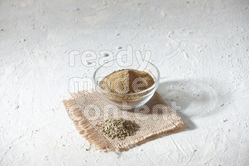 A glass bowl full of cumin powder with some of cumin seeds on burlap piece on a textured white flooring
