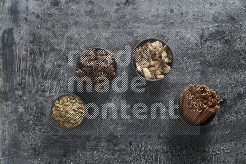 Ginger, Cardamom, Star anise and cinnamon sticks in 4 bowls on a textured black background