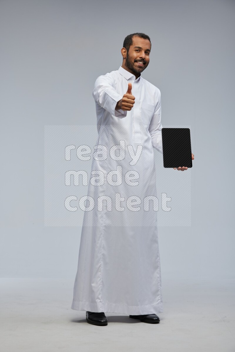 Saudi man wearing Thob standing showing tablet to camera on Gray background