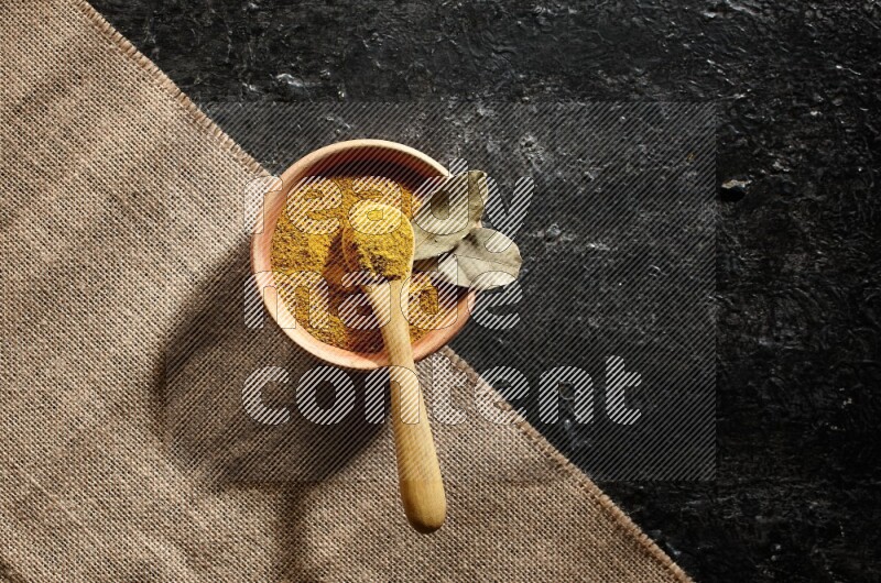 A wooden bowl and a wooden spoon full of turmeric powder on burlap fabric on textured black flooring