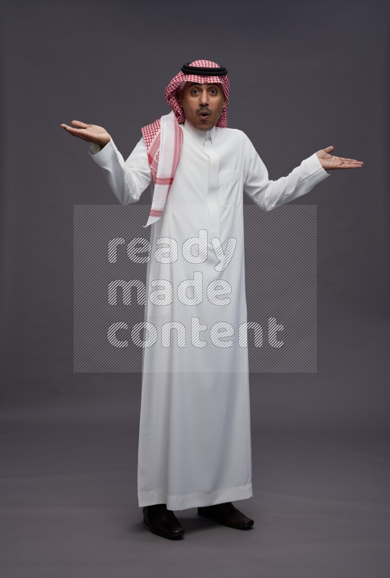 Saudi man wearing thob and shomag standing interacting with the camera on gray background