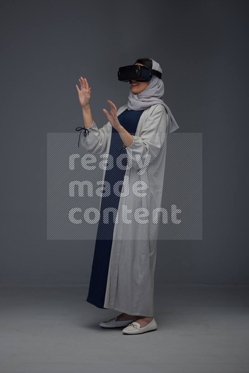 A Saudi woman wearing a light gray Abaya and head scarf standing and wearing VR eye level on a grey background