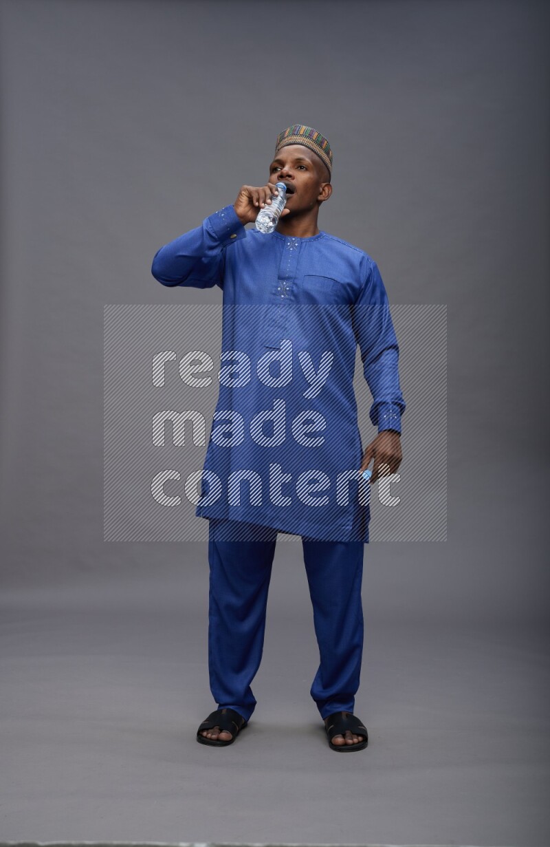 Man wearing Nigerian outfit standing drinking water on gray background