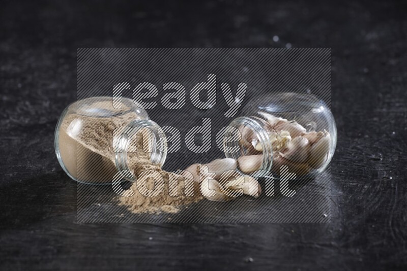 2 glass spice jars, one full of garlic powder and the other full of garlic cloves both flipped on a textured black flooring