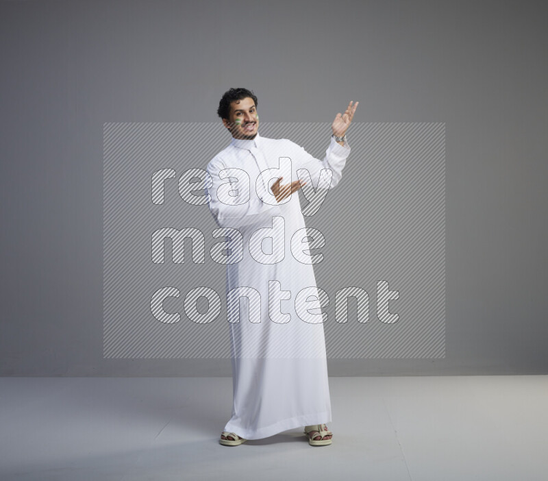 A Saudi man standing wearing thob with face painting interacting with the camera on gray background